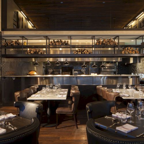 A modern restaurant with elegant dining tables, leather chairs, and an open kitchen stocked with firewood and cooking equipment, dimly lit ambiance.
