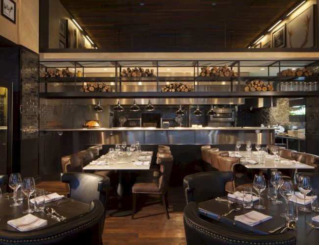 A modern restaurant with elegant dining tables, leather chairs, and an open kitchen stocked with firewood and cooking equipment, dimly lit ambiance.